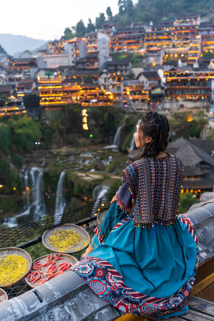 Viajes culturales a China: mujer con vestimenta tradicional mirando un pueblo antiguo iluminado con cascadas.